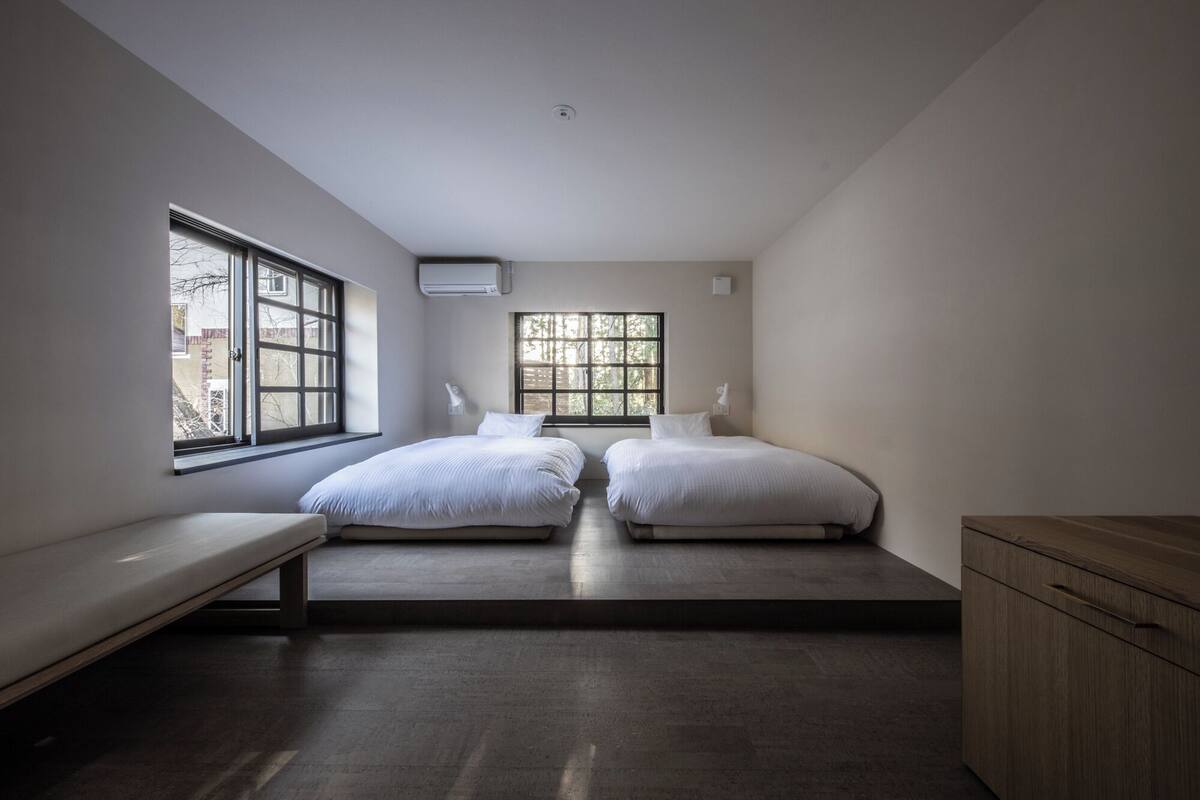 Un dormitorio con dos camas, un banco, un armario de madera y ventanas grandes.