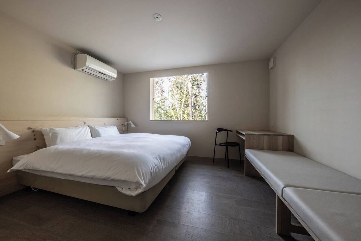 Un dormitorio con cama, banca, escritorio y ventana con vista a árboles.