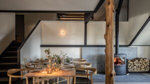 Un comedor moderno con mesas y sillas de madera, un arreglo floral central y un estufa de leña al fondo.