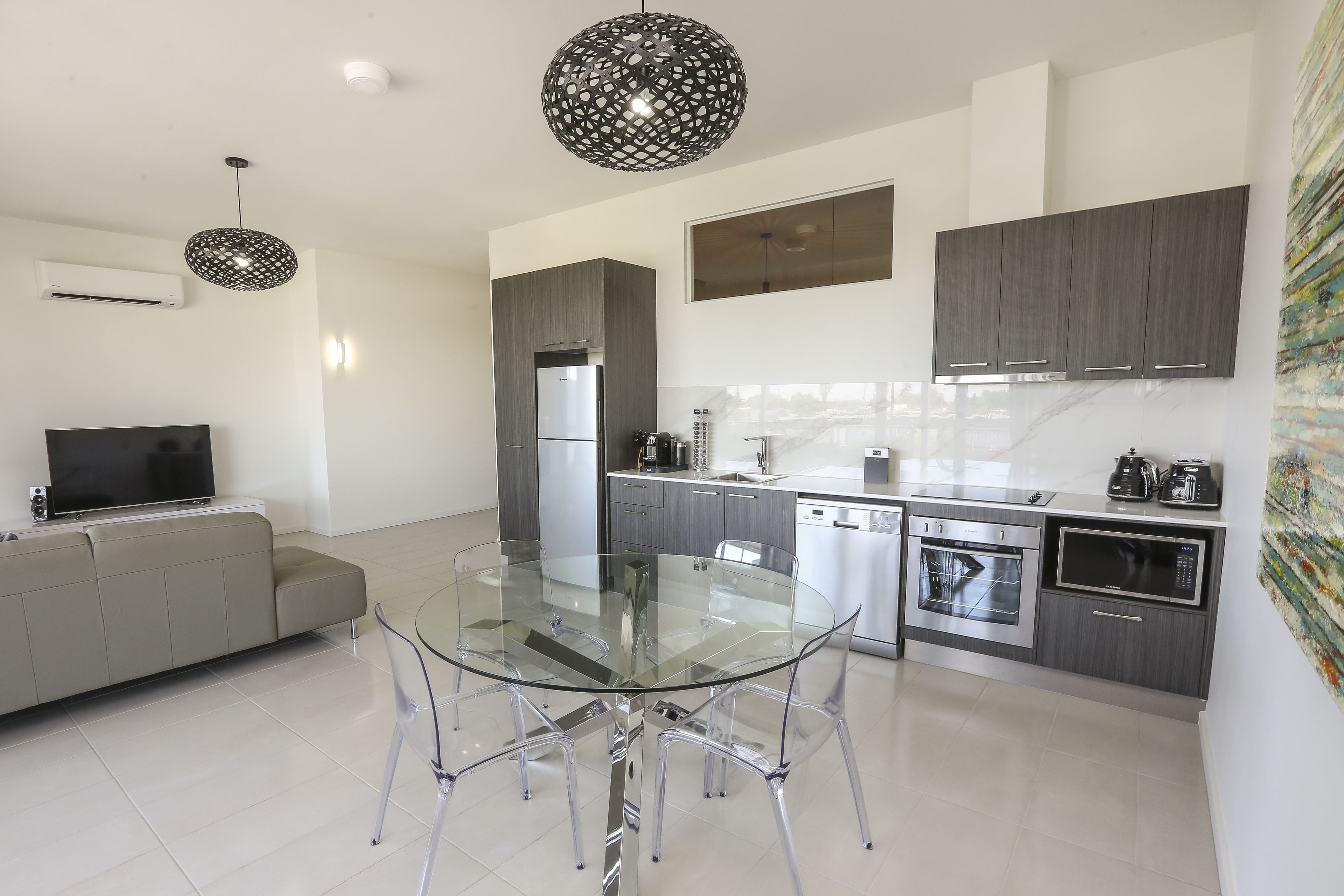A modern kitchen with a glass dining table, grey cabinets, stainless steel appliances, and a grey sofa.