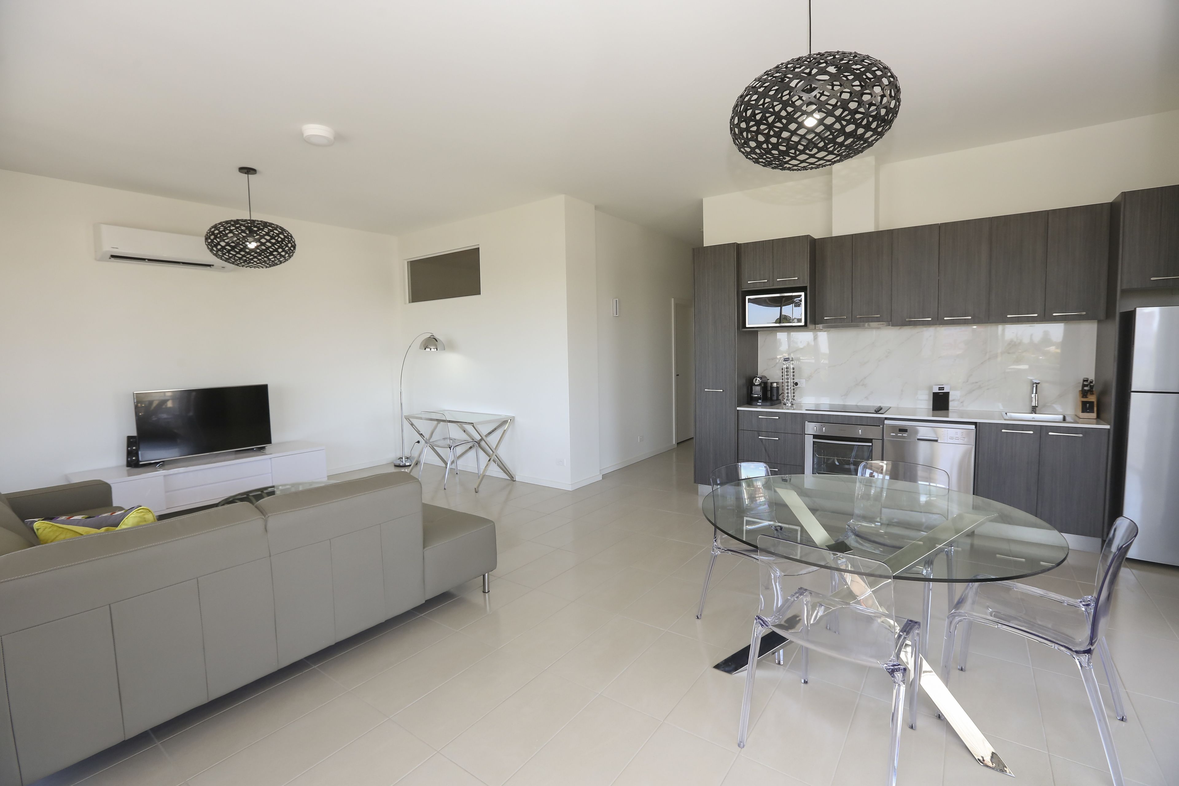 Un espacio habitable moderno con una mesa de comedor de vidrio, un sofá, un televisor y una cocina con armarios grises y electrodomésticos de acero inoxidable.