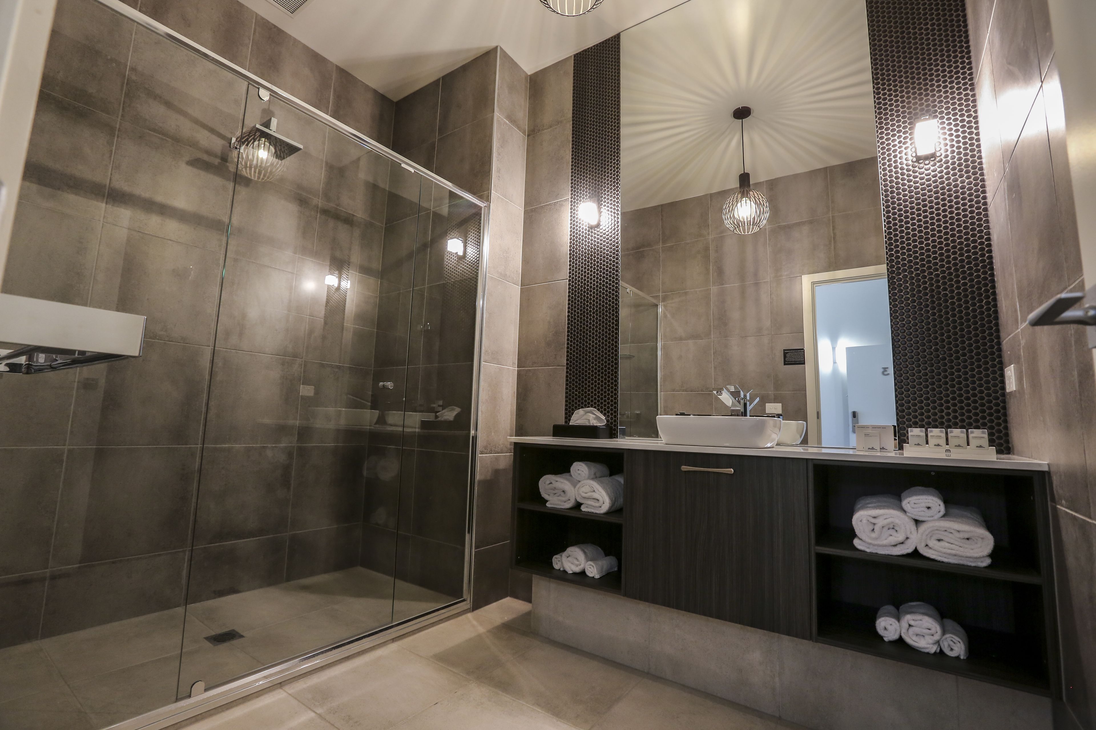A modern bathroom with a glass-enclosed shower, dark tiled walls, a large mirror, and shelves with neatly folded towels.