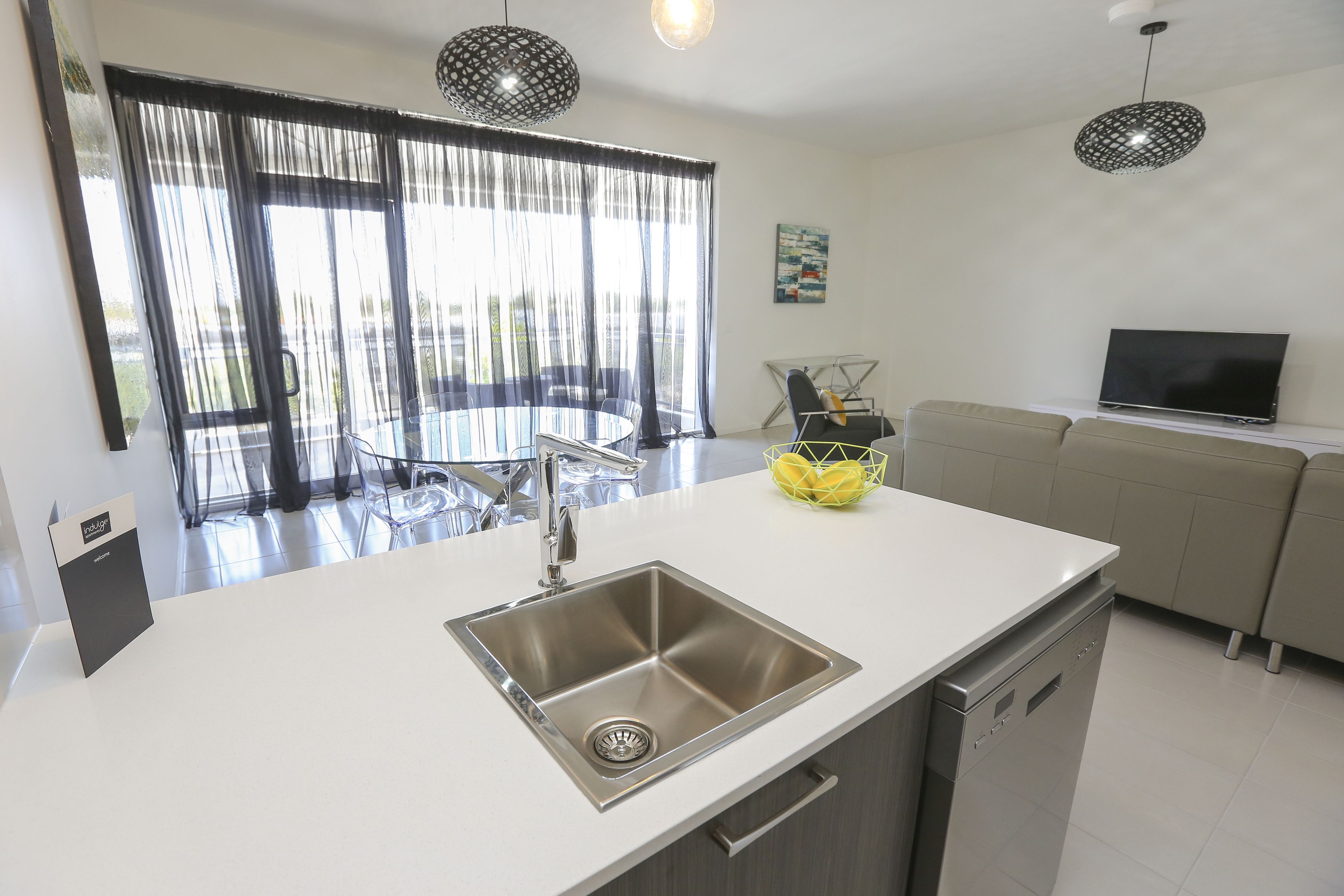 Una cocina moderna con un lavamanos de acero inoxidable, lavavajillas y un comedor con mesa y sillas de vidrio.