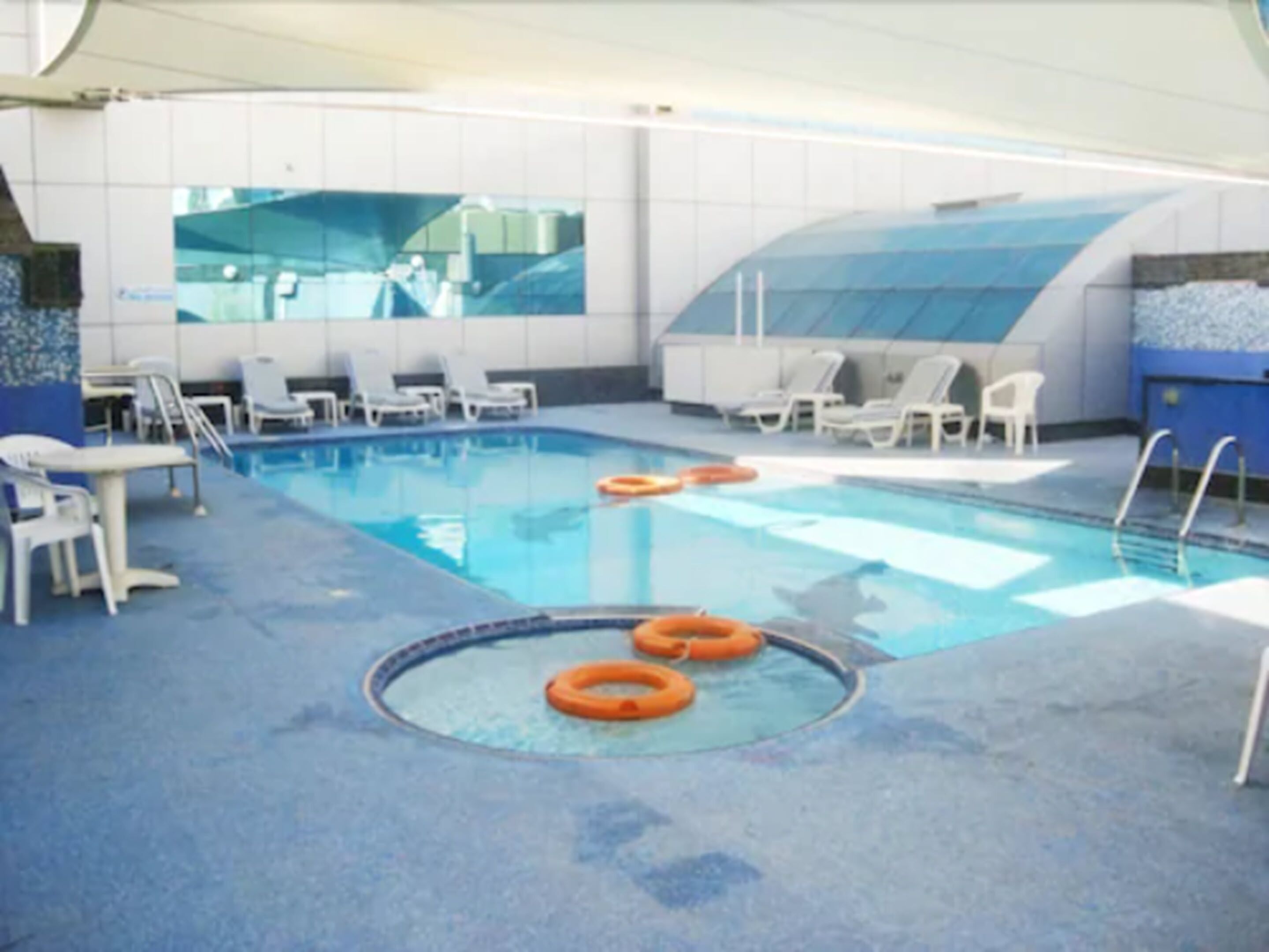 An indoor swimming pool with clear water, surrounded by white lounge chairs and orange life rings.