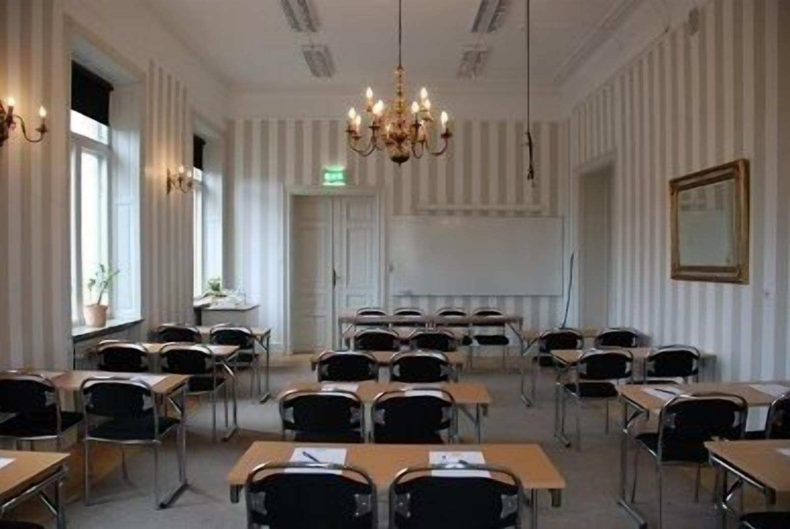 Ett konferensrum med rader av bord och stolar, en kristallkrona och en stor spegel på väggen.