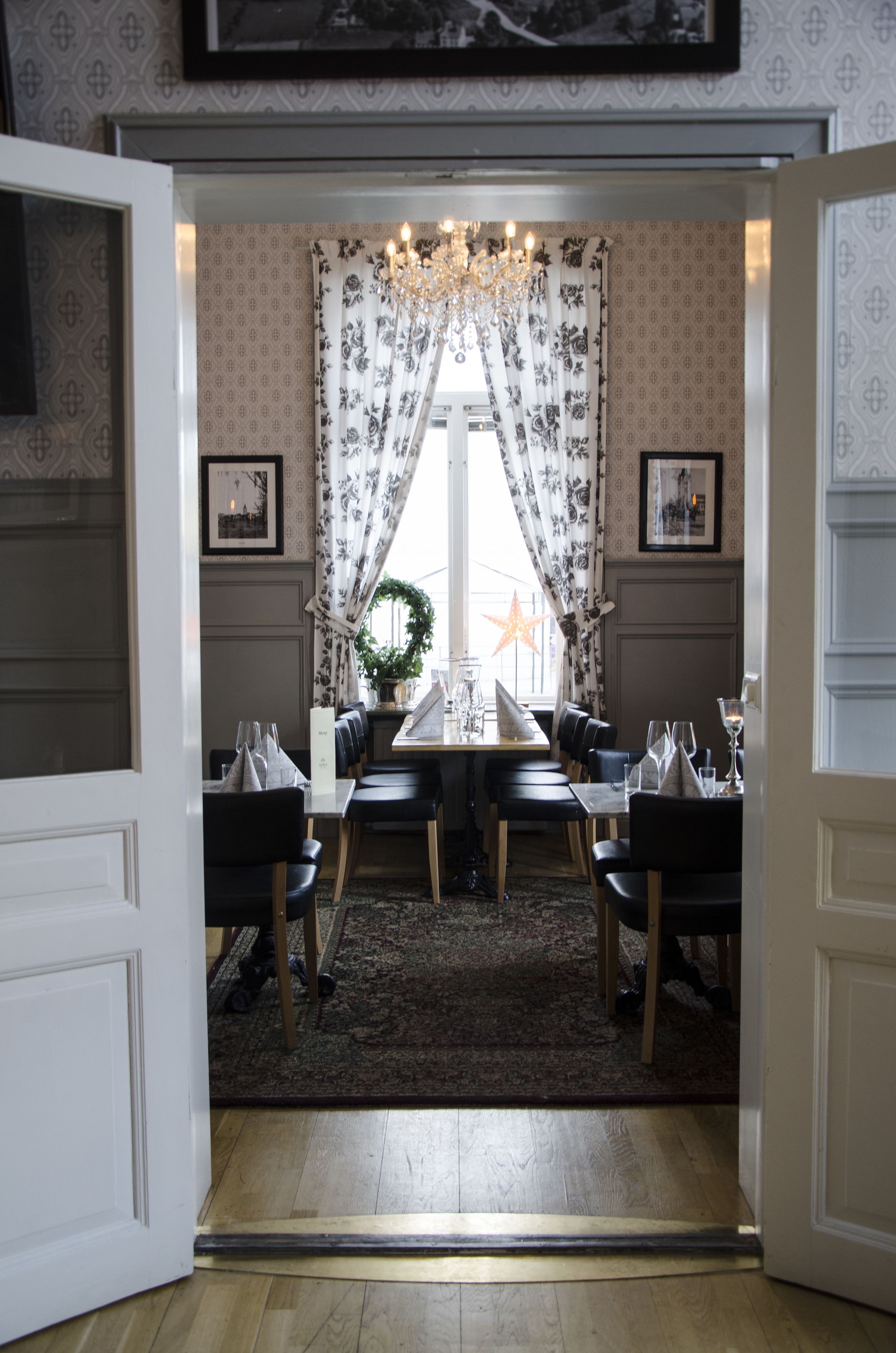 En matplats med ett bord dukat för fyra personer, omgiven av stolar, en kristallkrona och ett fönster med ett blommönster och gardiner.