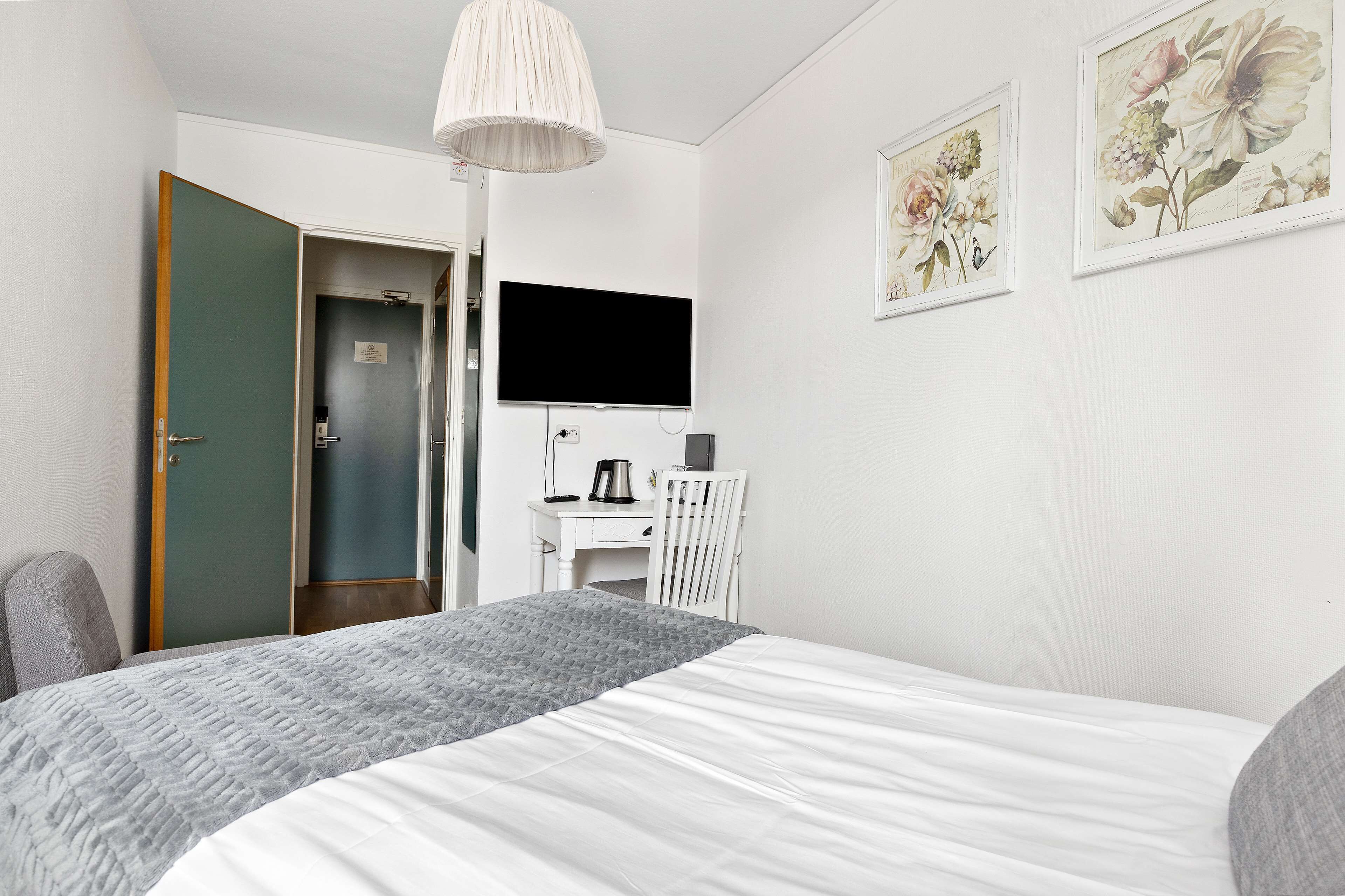 Ett sovrum med en säng, ett skrivbord, en stol, en tv och två inramade konstverk med blommotiv på väggen.