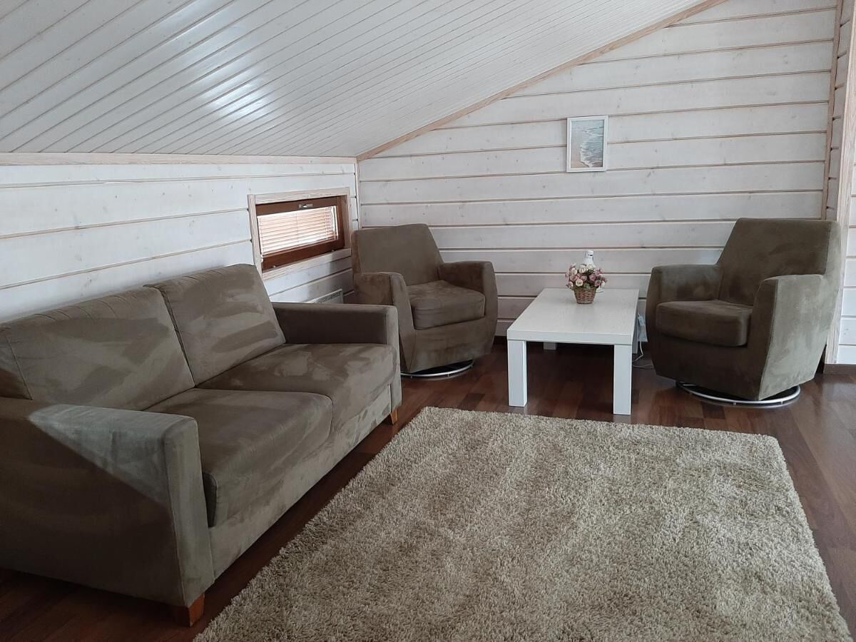 A cozy attic room with a wooden sloped ceiling, a brown sofa, two armchairs, a small white table, and a beige rug.
