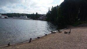 Escena junto a un lago con un bote amarrado en la orilla, rodeado de árboles y un edificio al fondo.