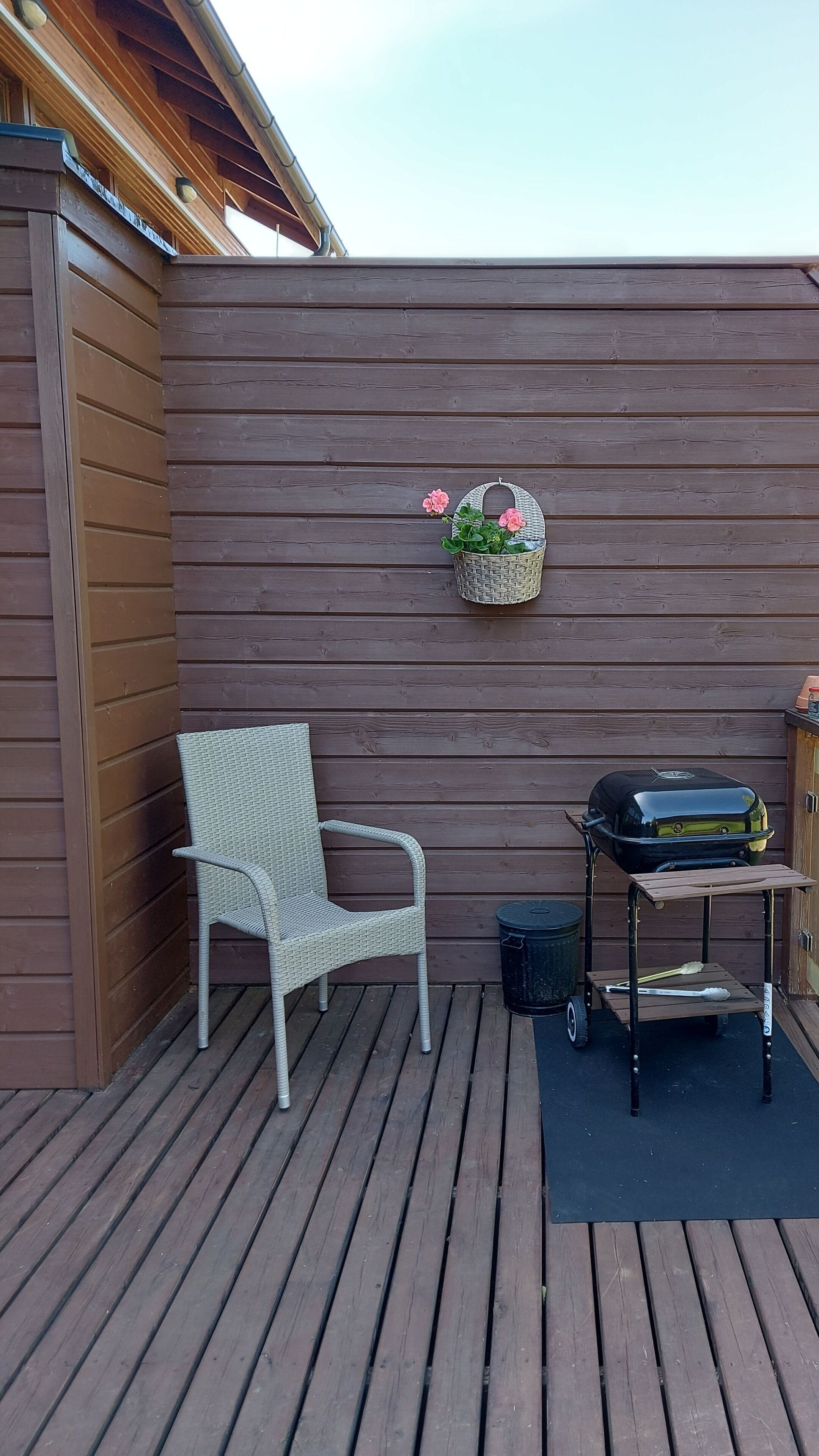 A wooden deck with a wicker chair, a grill cart, and a hanging basket with flowers.