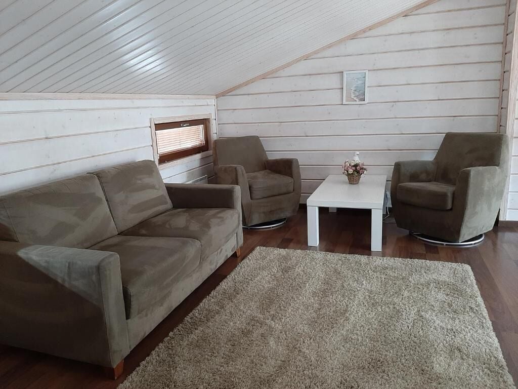 Um quarto acolhedor em estilo loft, com teto alto, paredes de madeira, um sofá bege, duas poltronas, uma mesa pequena branca e um tapete.