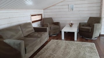 A cozy loft-style room with a high ceiling, wooden walls, a beige sofa, two armchairs, a small white table, and a rug.