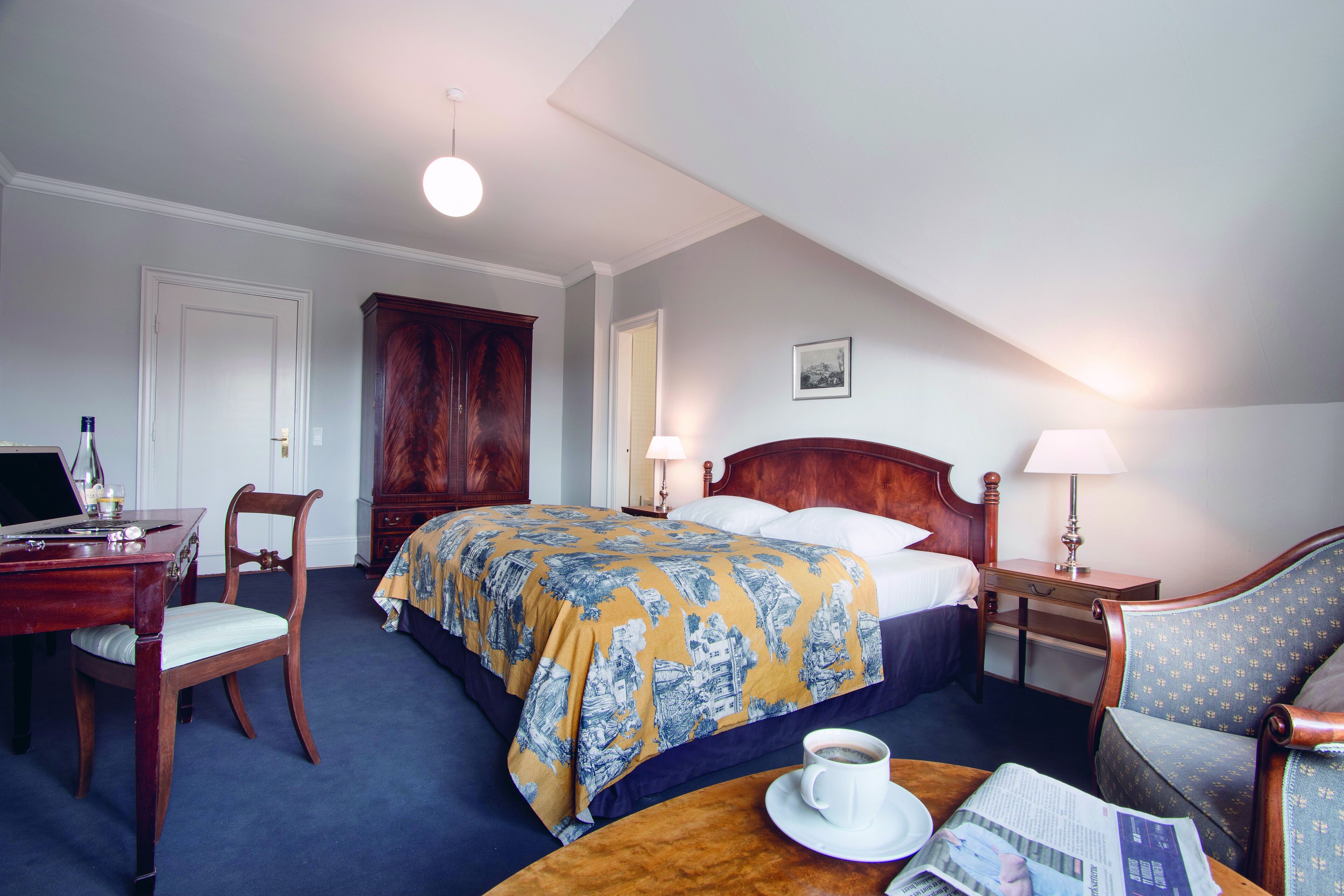 Et hotelværelse med en seng, et skrivebord med en bærbar computer, en stol, et sofabord med en kop og en avis, samt en lampe.