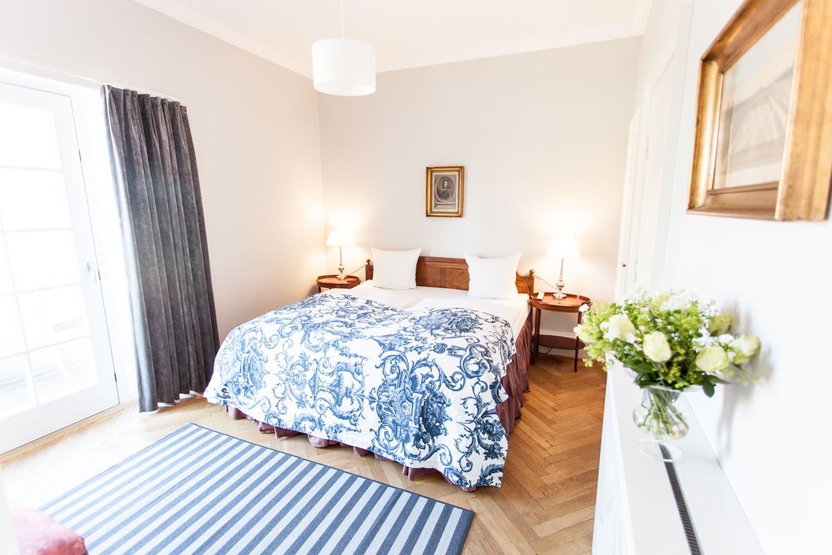 A hotel room with a large bed, two bedside tables with lamps, a framed picture on the wall, a window with curtains, and a vase with flowers on a nightstand.