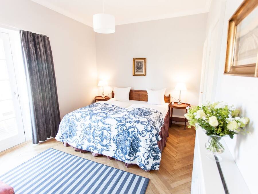 Een hotelkamer met een groot bed, twee nachtkastjes met lampen, een lijst met een afbeelding aan de muur, een raam met gordijnen en een vaas met bloemen op een nachtkastje.