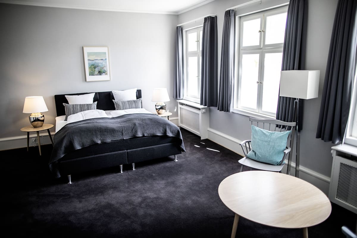 A modern hotel room with a large bed, two bedside tables with lamps, a small round table, and a window with curtains.