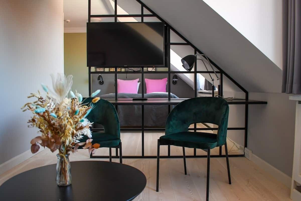 A modern bedroom with a sloped ceiling, a bed with pink and gray bedding, two green chairs, a black table with a vase of dried flowers, and a black TV mounted on the wall.