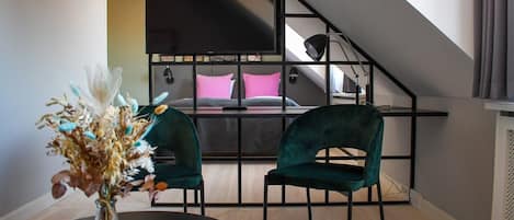 Un dormitorio moderno con techo inclinado, una cama con ropa de cama rosa y gris, dos sillas verdes, una mesa negra con un jarrón de flores secas y un televisor negro montado en la pared.