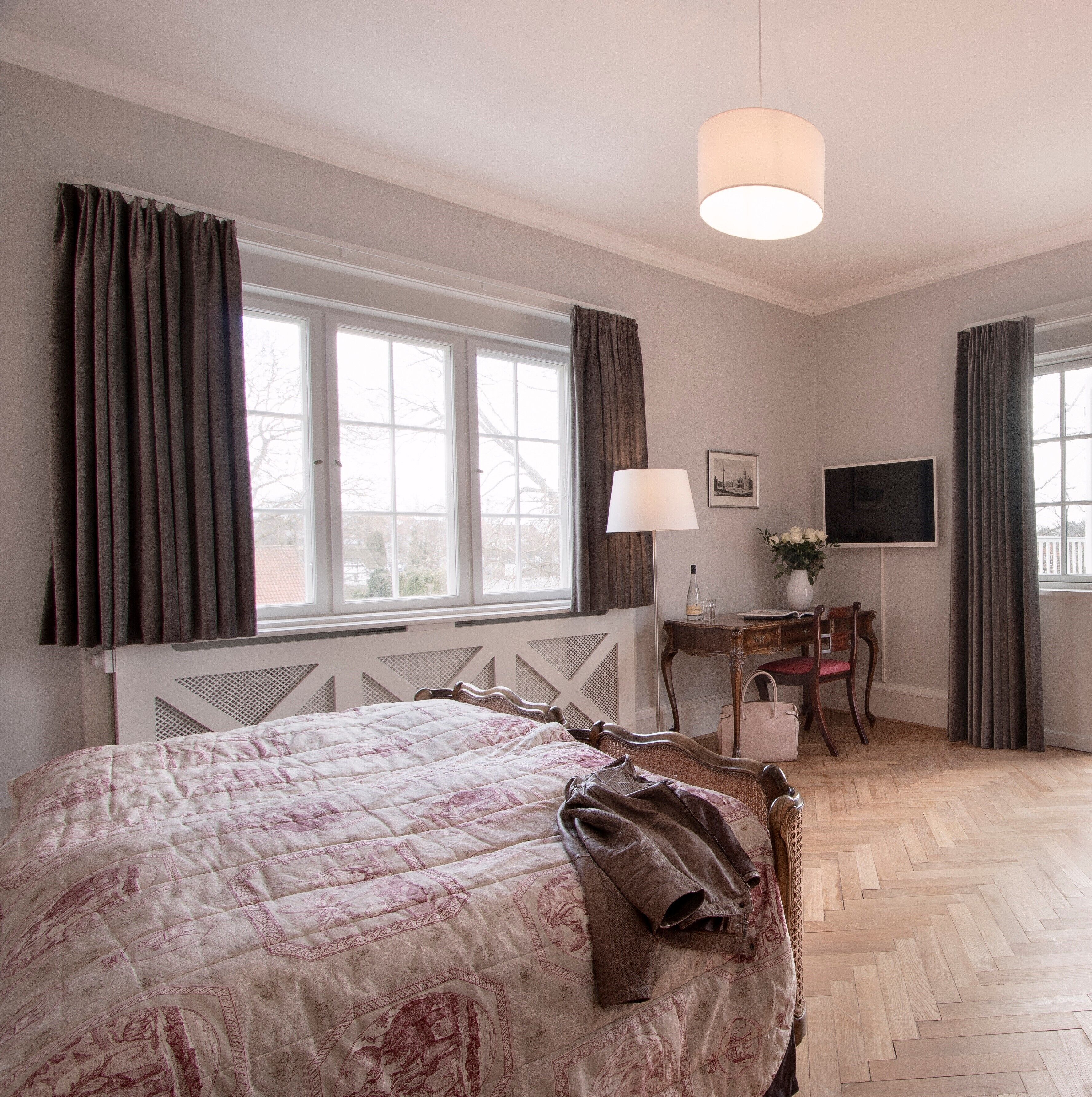 A bedroom with a large bed, a desk, a chair, a television, and a window with curtains.