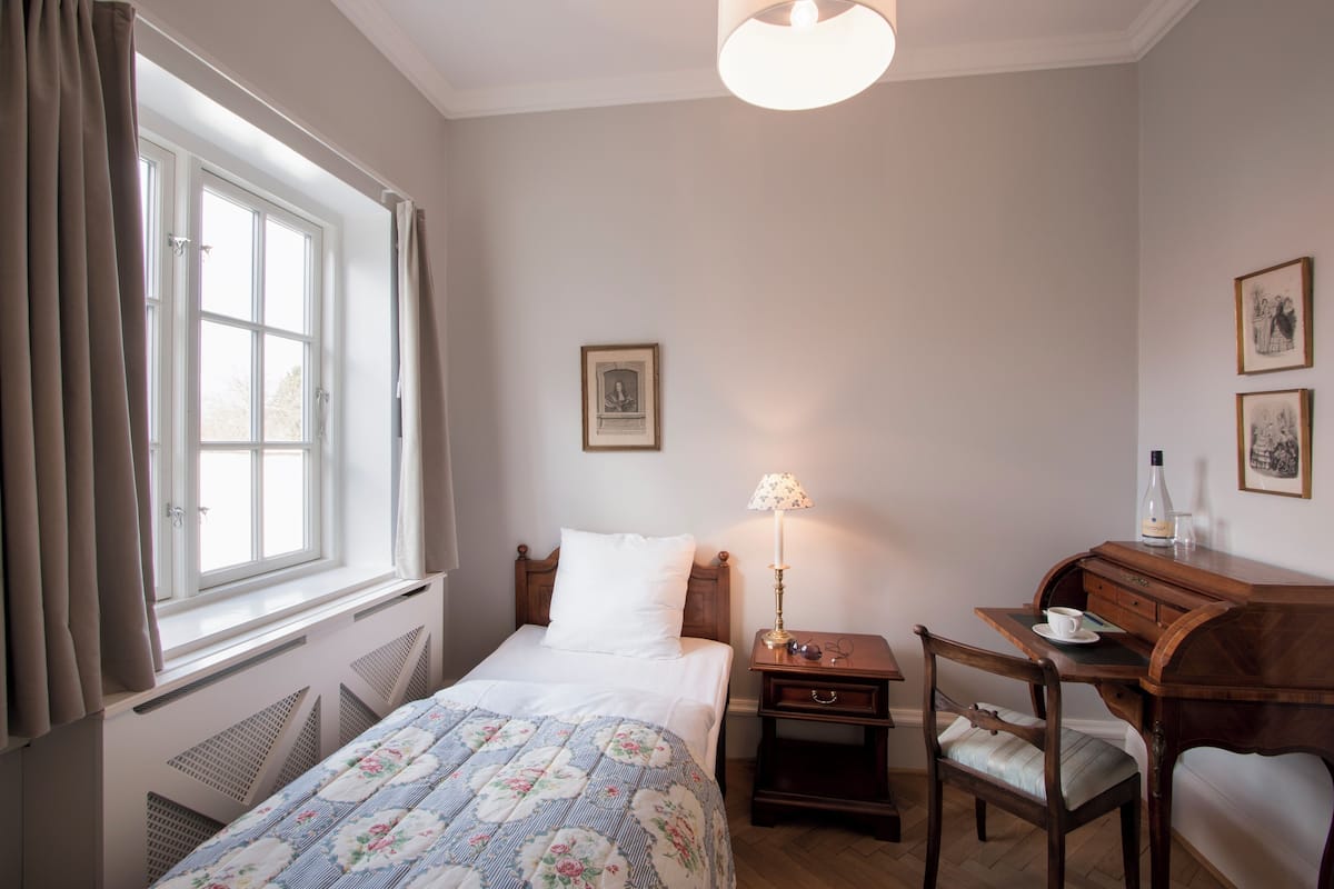 A bedroom with a bed, a desk, a chair, a lamp, and a window with curtains.