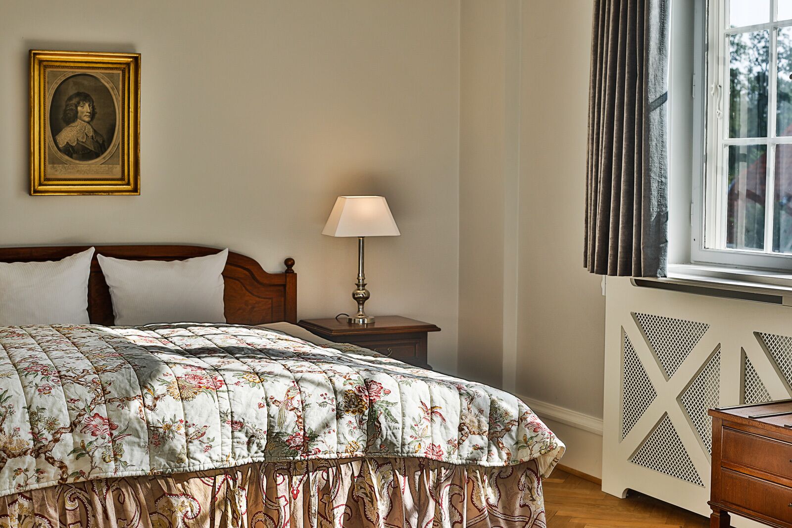 A bedroom with a wooden bed, a nightstand with a lamp, a framed portrait on the wall, and a window with curtains.
