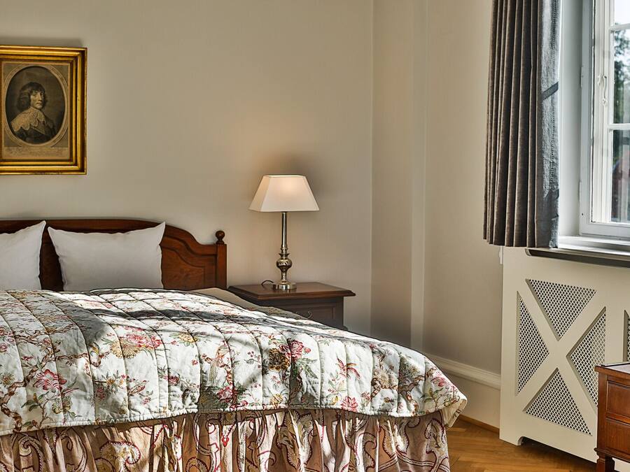 A bedroom with a wooden bed, a nightstand with a lamp, a framed portrait on the wall, and a window with curtains.