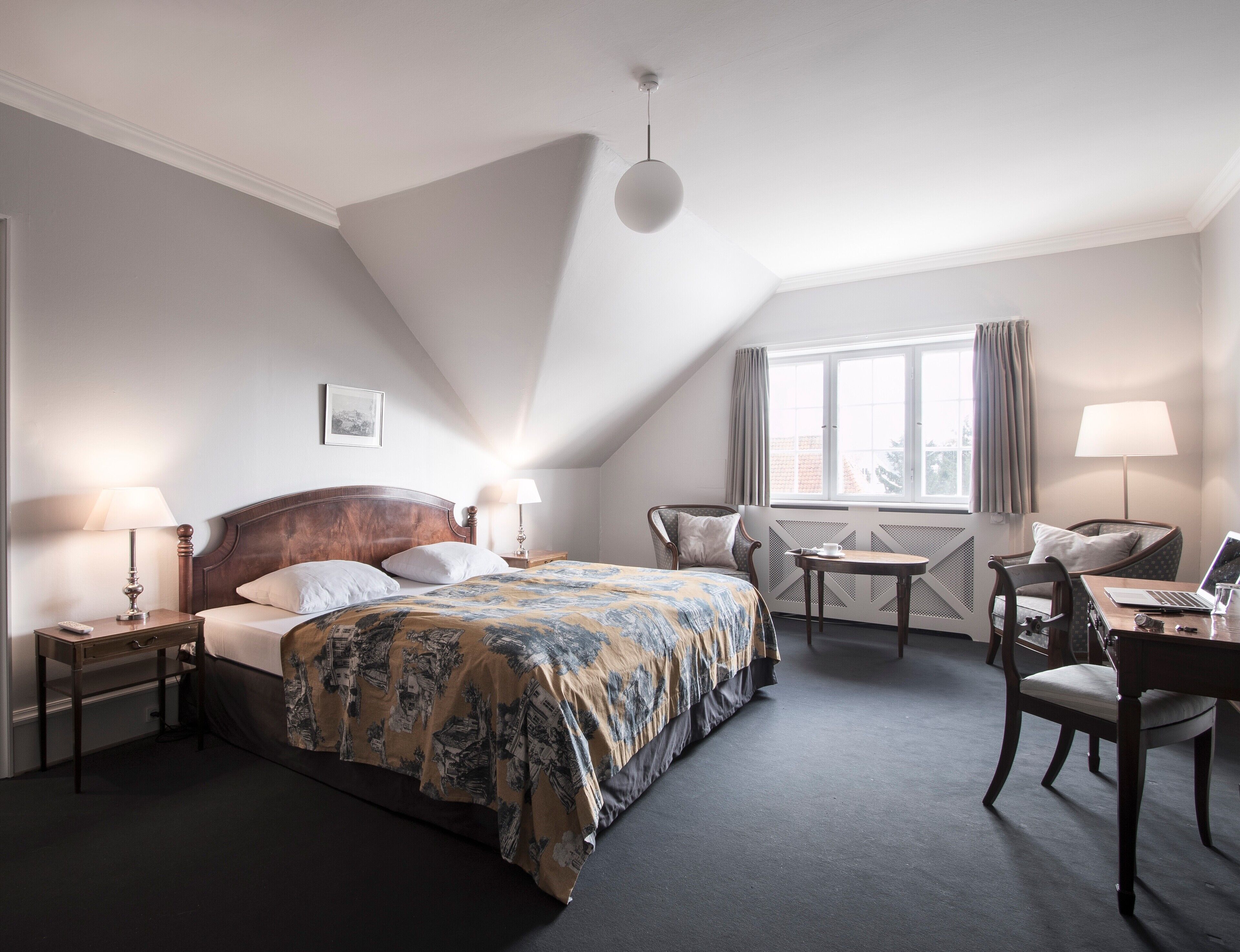 A hotel room with a bed, a desk with a laptop, a chair, a window with curtains, and a lamp on the nightstand.