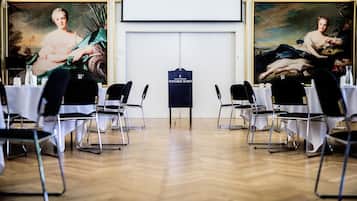 A conference room with tables and chairs, a projector screen, and two large classical paintings on the walls.