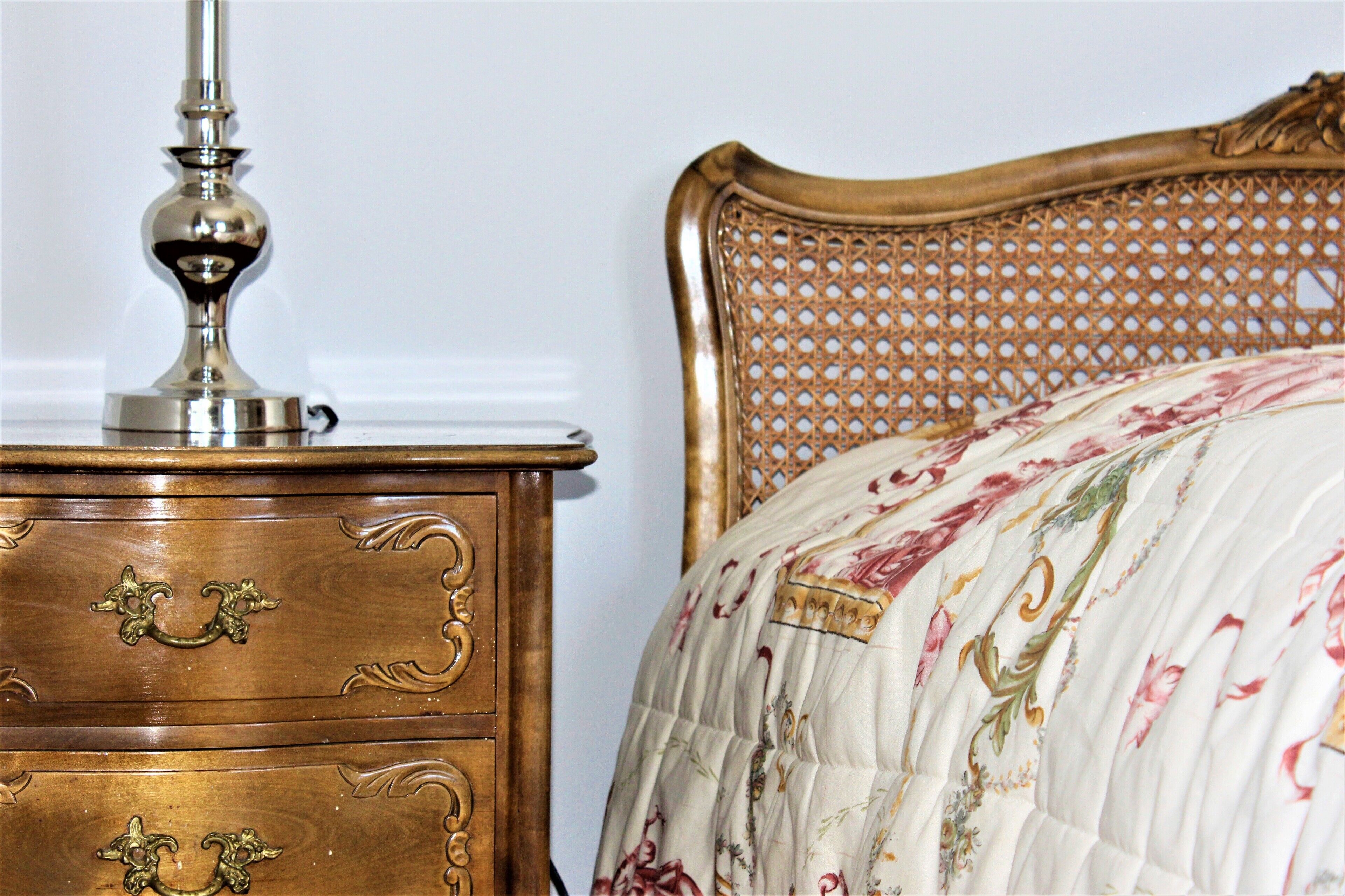 Un aparador de madera con adornos en los tiradores y una lámpara metálica, junto a una cama con colcha estampada de flores y cabecero tejido.