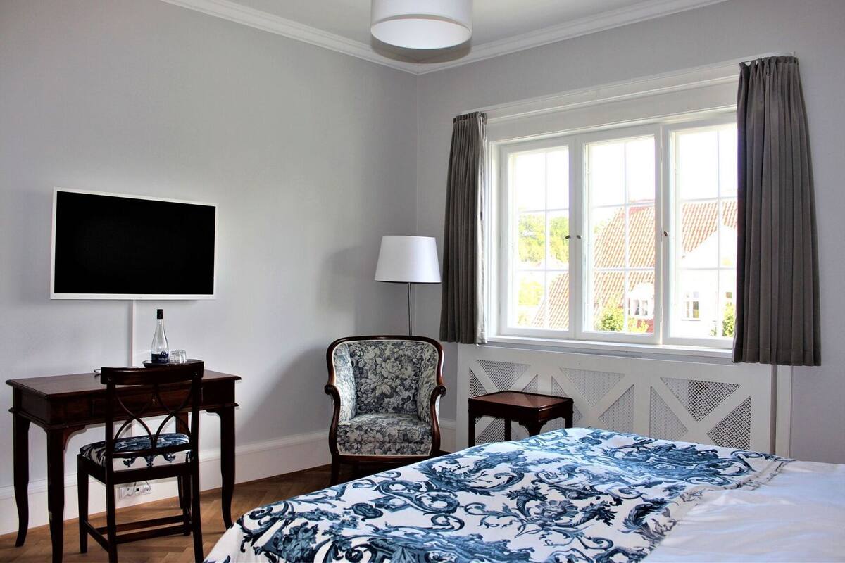 A hotel room with a bed, a desk, a chair, a TV, a window with curtains, and a lamp.