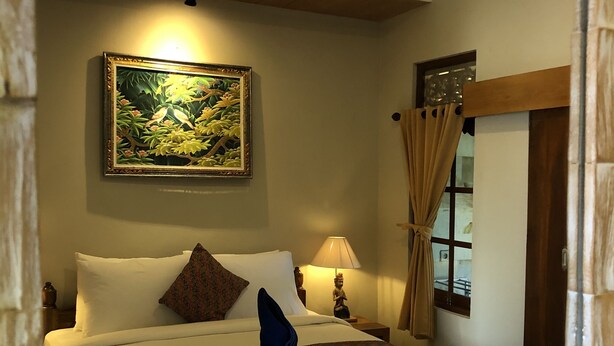A neatly made bed with white linens, a decorative pillow, and a blue folded item on the bed. There is a painting on the wall above the bed, a lamp on the nightstand, and a window with curtains. - Jangkrik Homestay (Ubud)