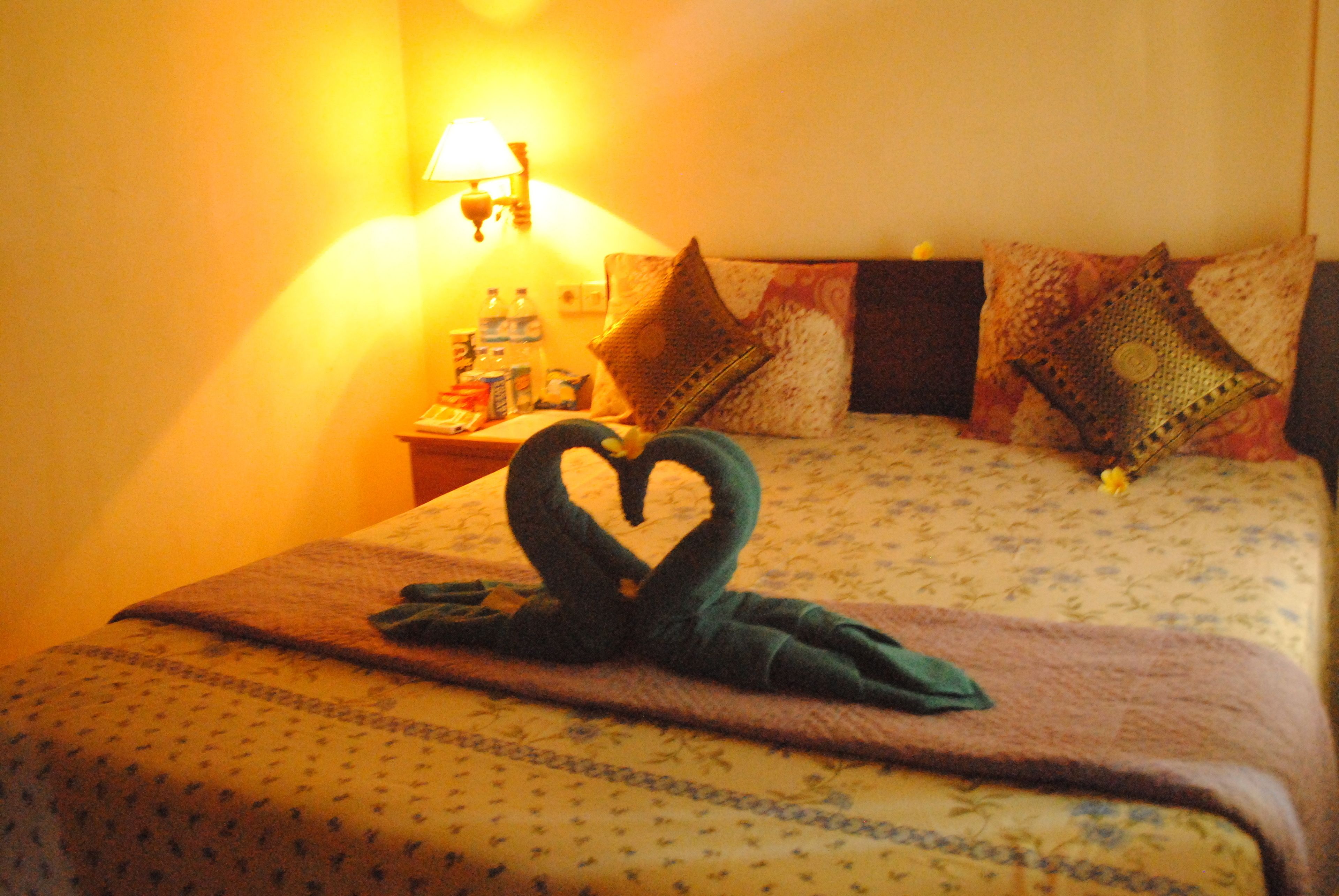 Quarto de hotel com cama, mesa de cabeceira com abajur e toalha em formato de coração sobre a cama.