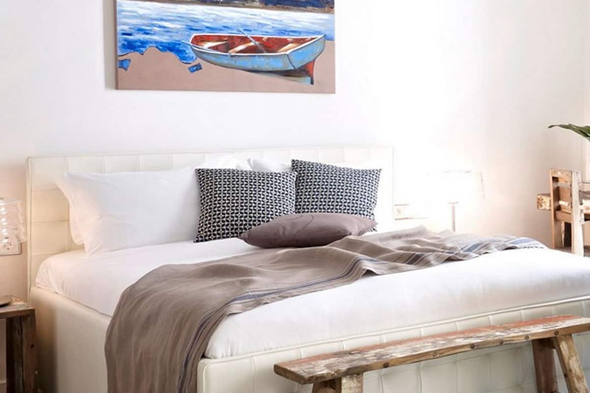 A bedroom with a bed, pillows, a blanket, a painting, a wooden bench, and a striped rug.