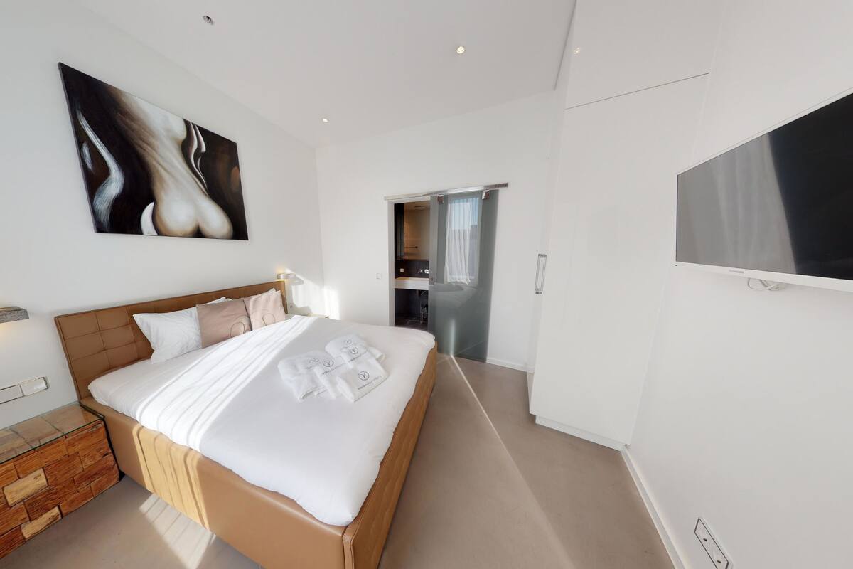 A modern hotel room with a large bed, a flat-screen TV, a painting above the bed, and a small wooden chest of drawers.