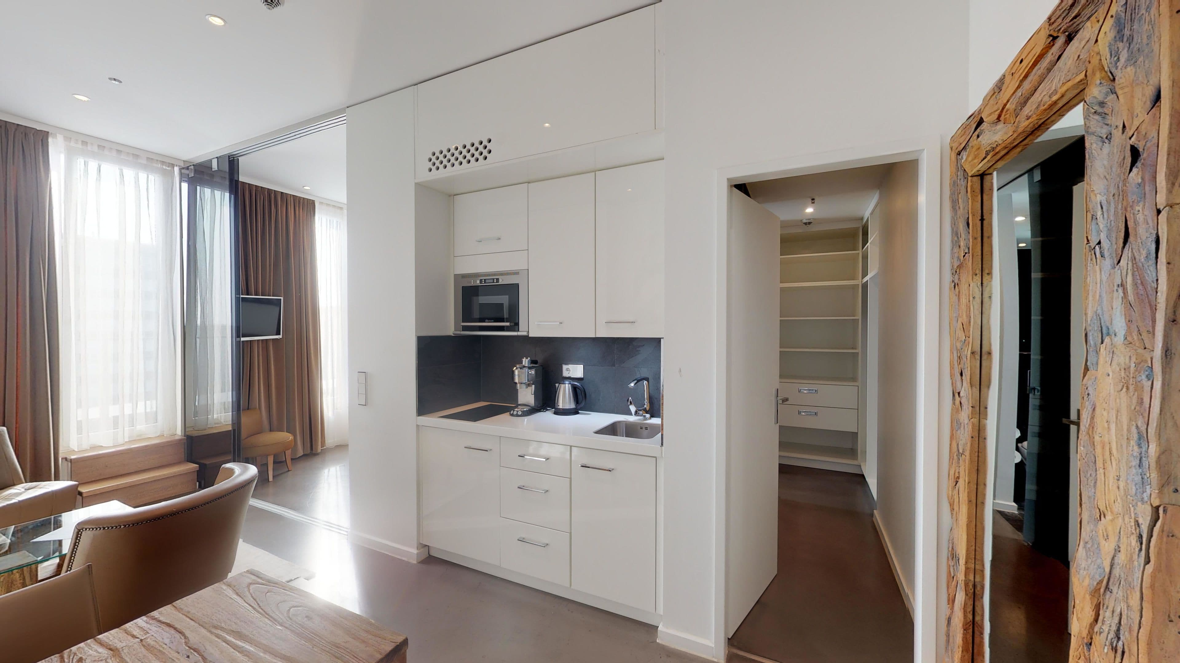 A modern kitchen with white cabinets, a built-in microwave, and a coffee maker. There is a dining area with a wooden table and chairs.