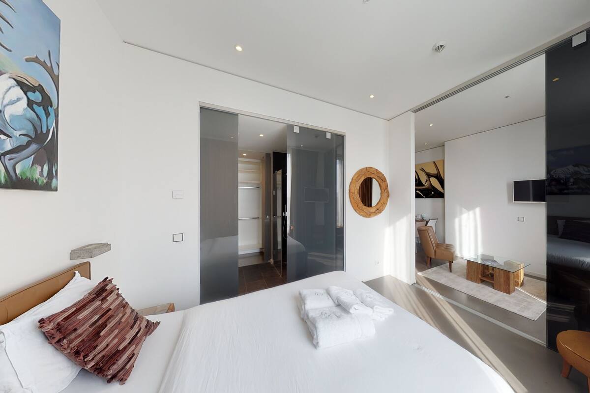 A modern hotel room with a large bed, a glass partition, a round mirror, a TV, and a painting on the wall.