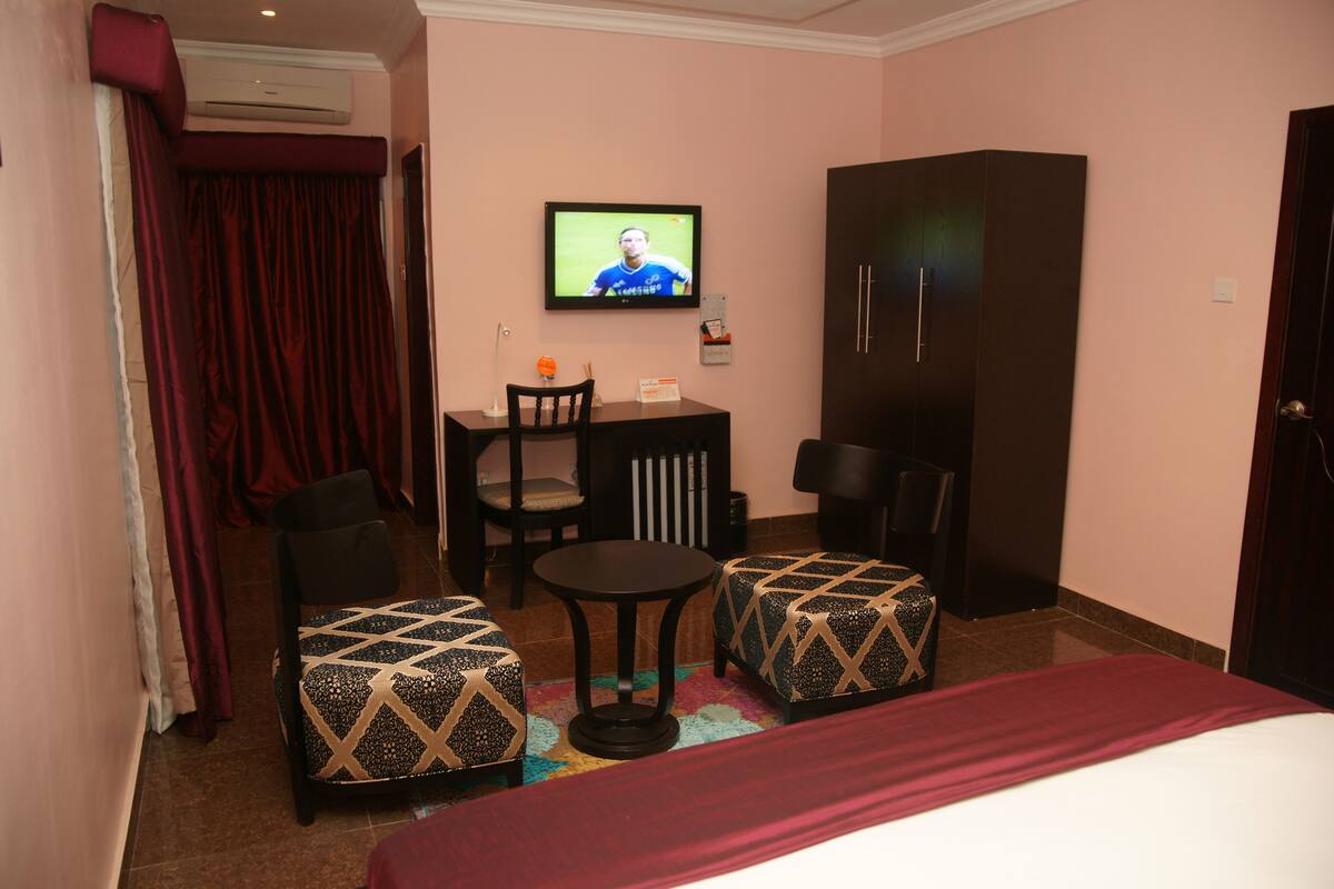 A hotel room with a bed, two patterned ottomans, a small table with chairs, a TV on the wall, and a wardrobe.