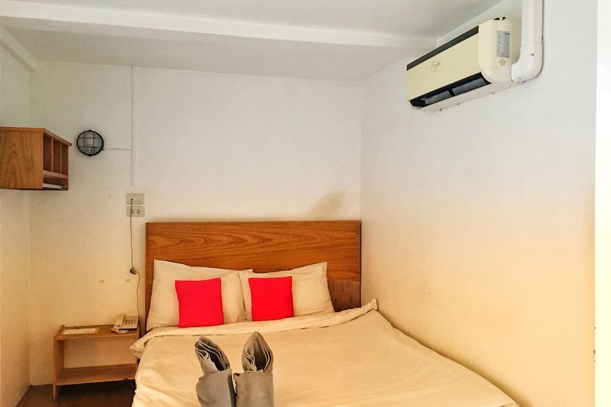 En säng för en person med en trägavel, två röda kuddar, en beige filt och ett par skor på golvet. Det finns ett litet träbord bredvid sängen med en telefon och en lampa. En luftkonditioneringsanläggning är monterad på väggen.