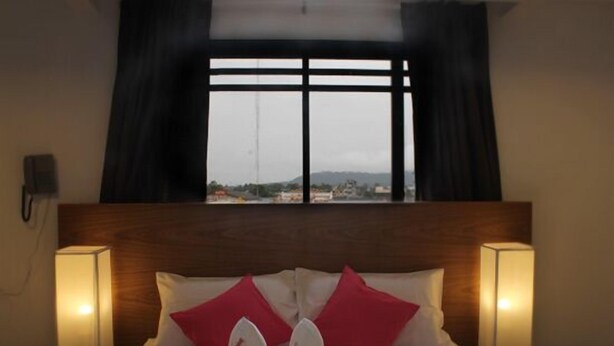 A hotel room with a bed, two bedside lamps, a window with curtains, and a view of a cityscape. - I Room Hotel (Surat Thani)