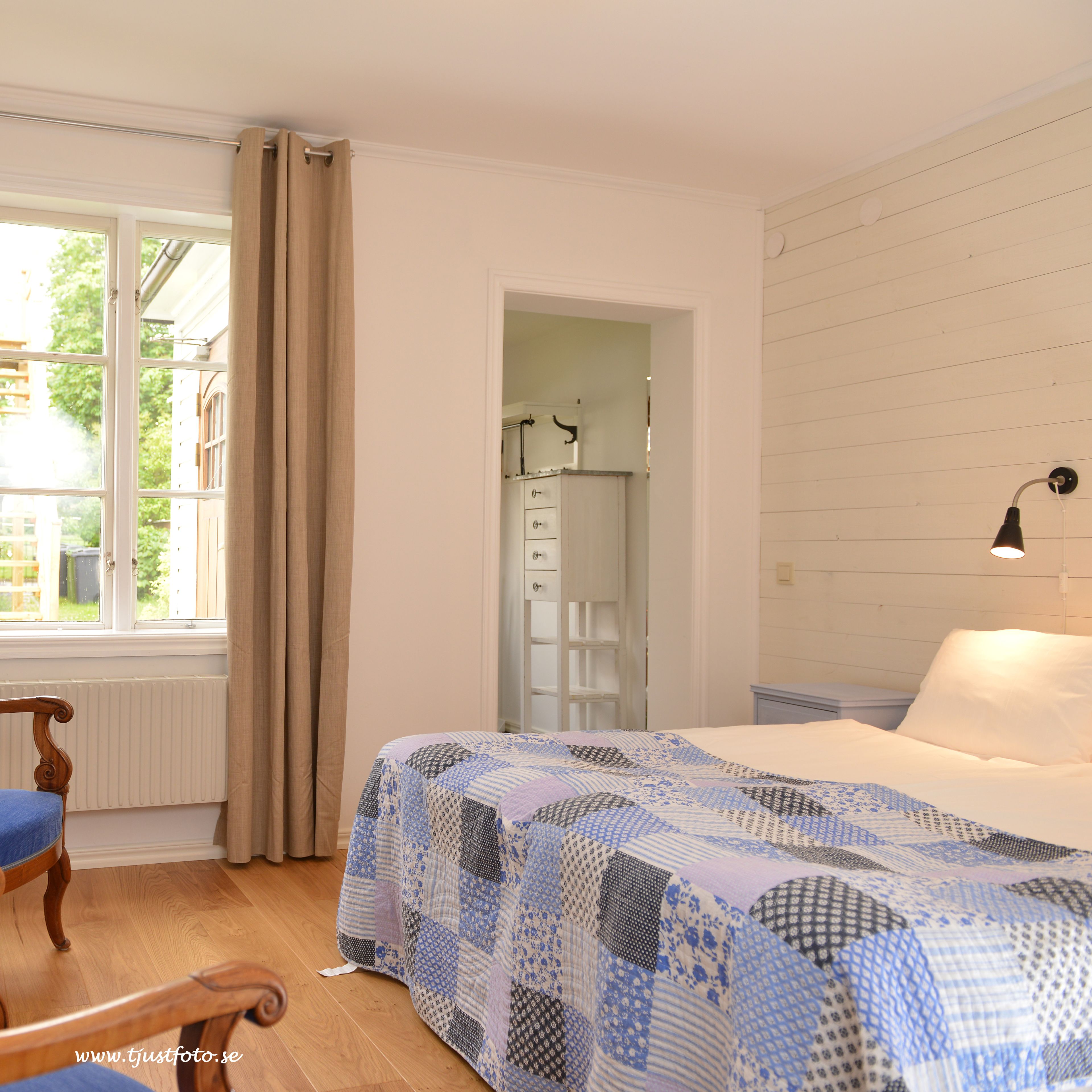 Un dormitorio con cama, una silla, una ventana con cortinas y un baño visible a través de una puerta abierta.