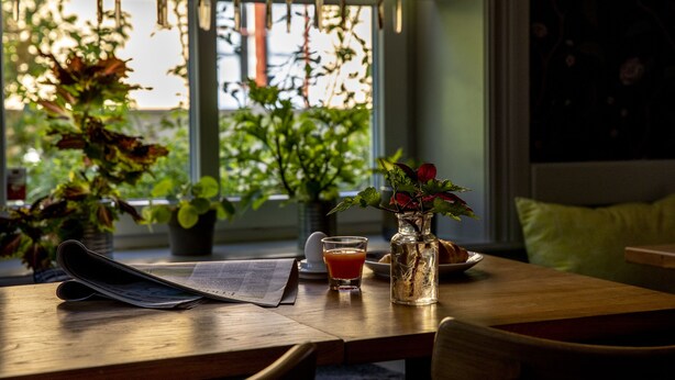 Une table en bois, un journal, un verre de jus d’orange, un vase avec des fleurs et une assiette contenant un croissant.