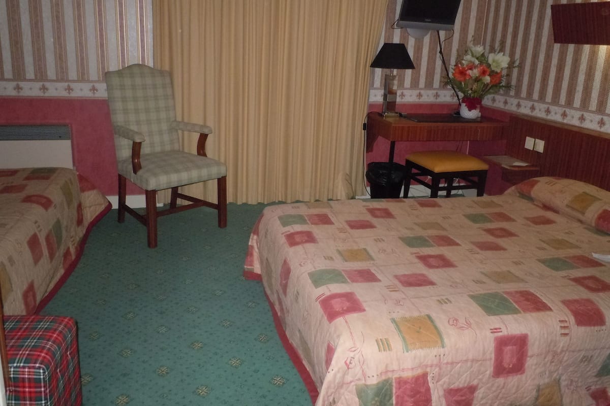 A hotel room with two beds, a chair, a desk with a lamp, a TV, and a small table with a vase of flowers.