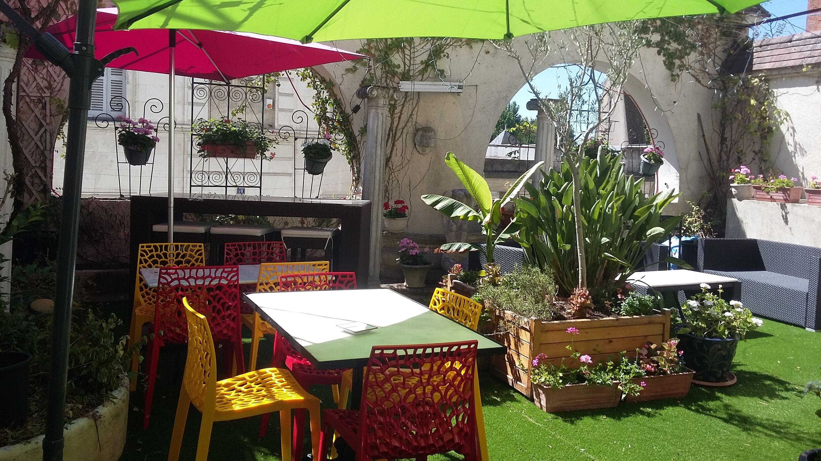 An outdoor patio with tables and chairs, surrounded by potted plants and a green lawn.