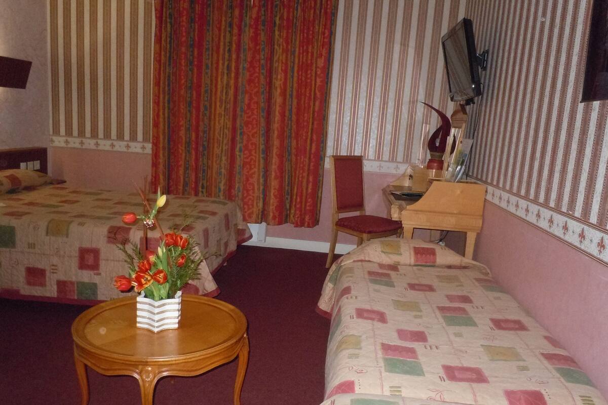A hotel room with two beds, a wooden table with a vase of flowers, a chair, a desk with a lamp, and a mirror on the wall.