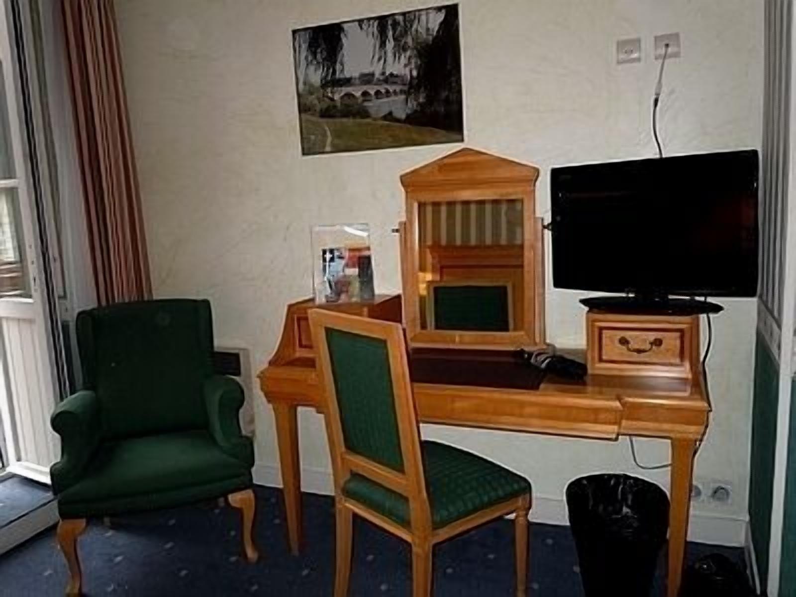 A room with a wooden desk, a green armchair, a television, and a picture of a bridge on the wall.