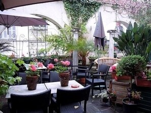 Un patio al aire libre con mesas y sillas, rodeado de plantas en macetas y una pérgola.