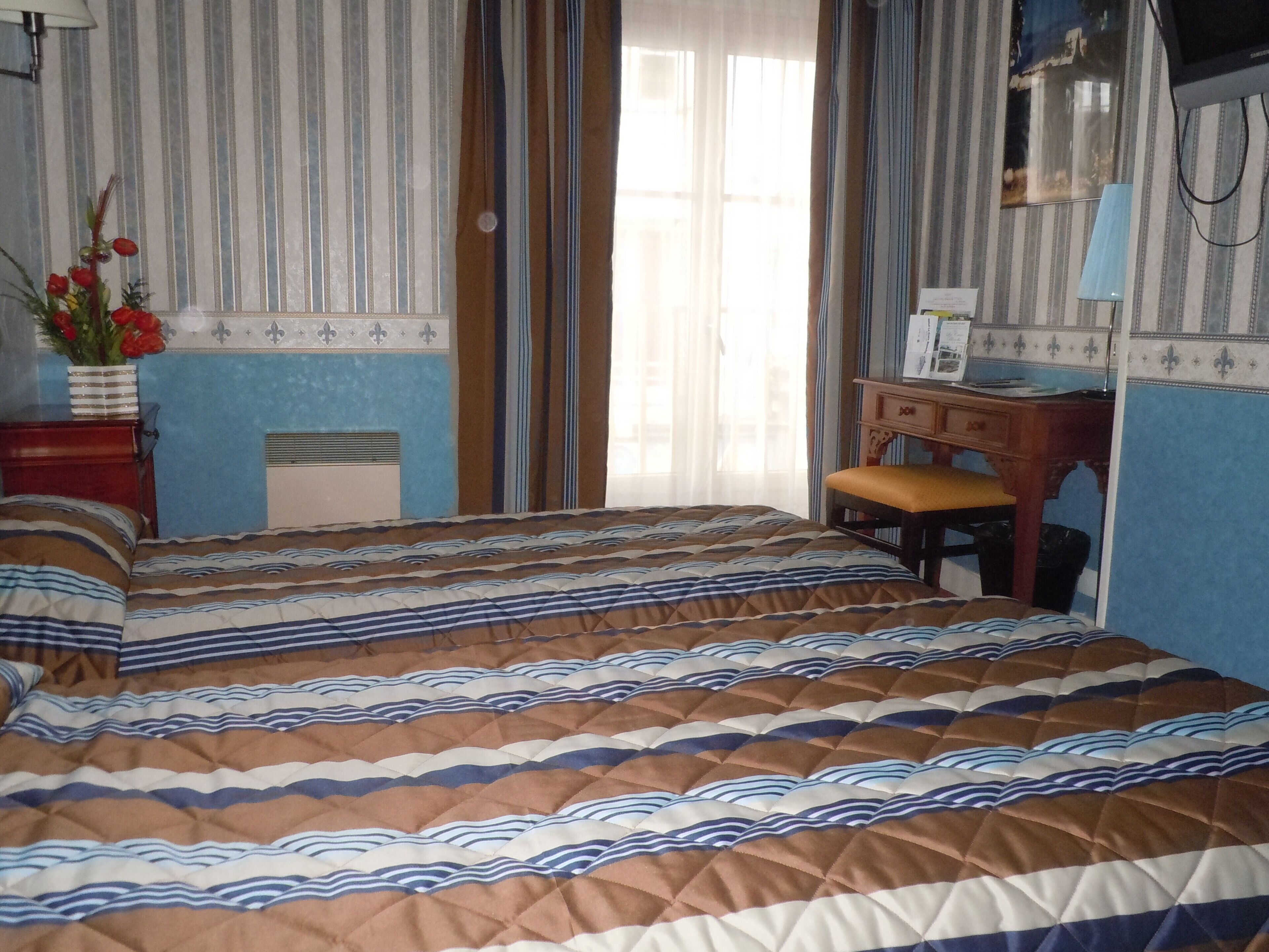 A bedroom with a bed, a desk, and a chair. There is a window with curtains, a radiator, and a vase with flowers on the nightstand.