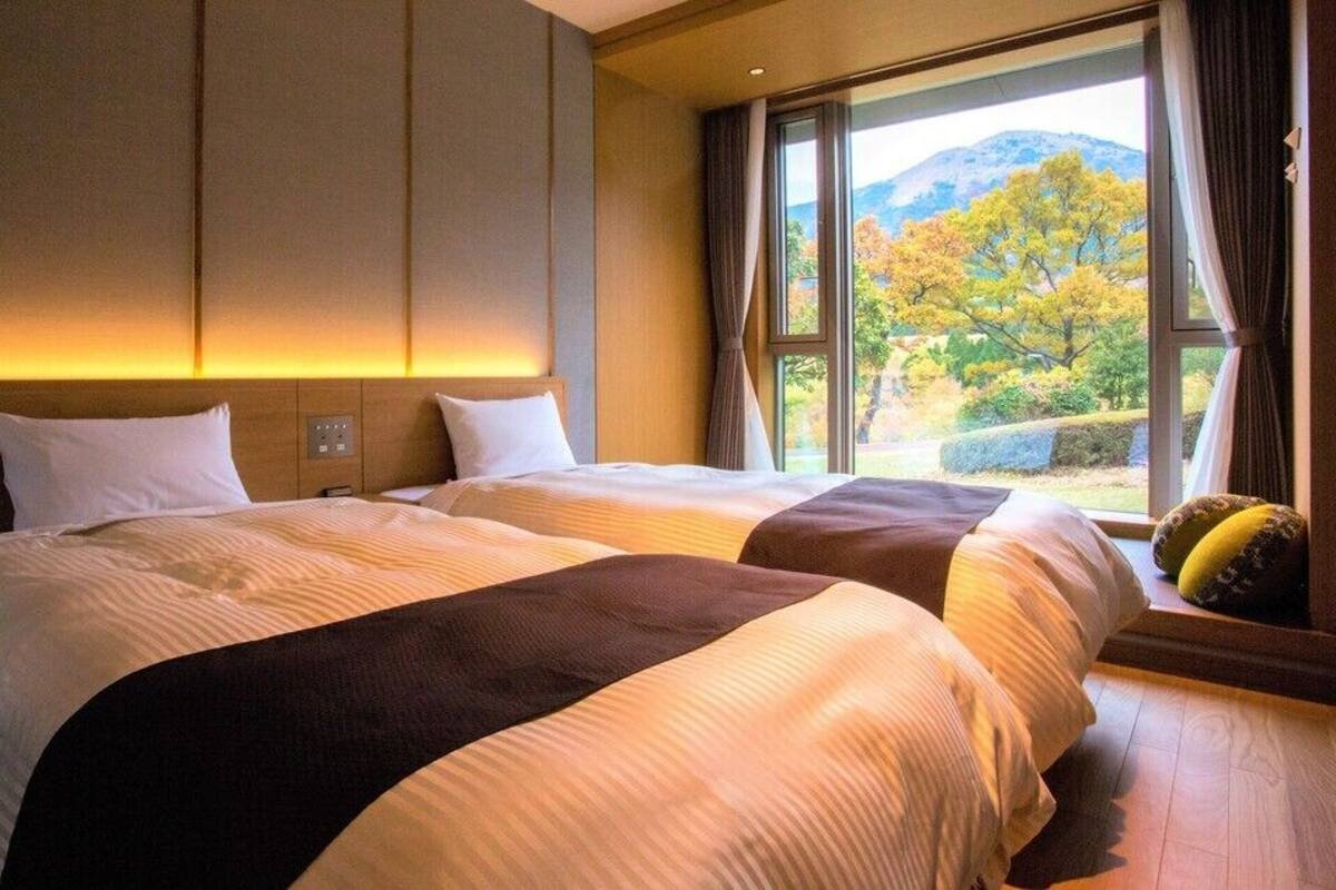 A hotel room with two beds, a large window with a view of trees and mountains, and a wooden floor.