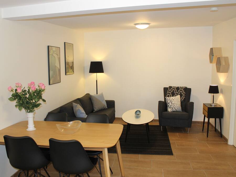 A modern living room with a wooden dining table, two black chairs, a grey sofa, a black armchair, and a small white table.