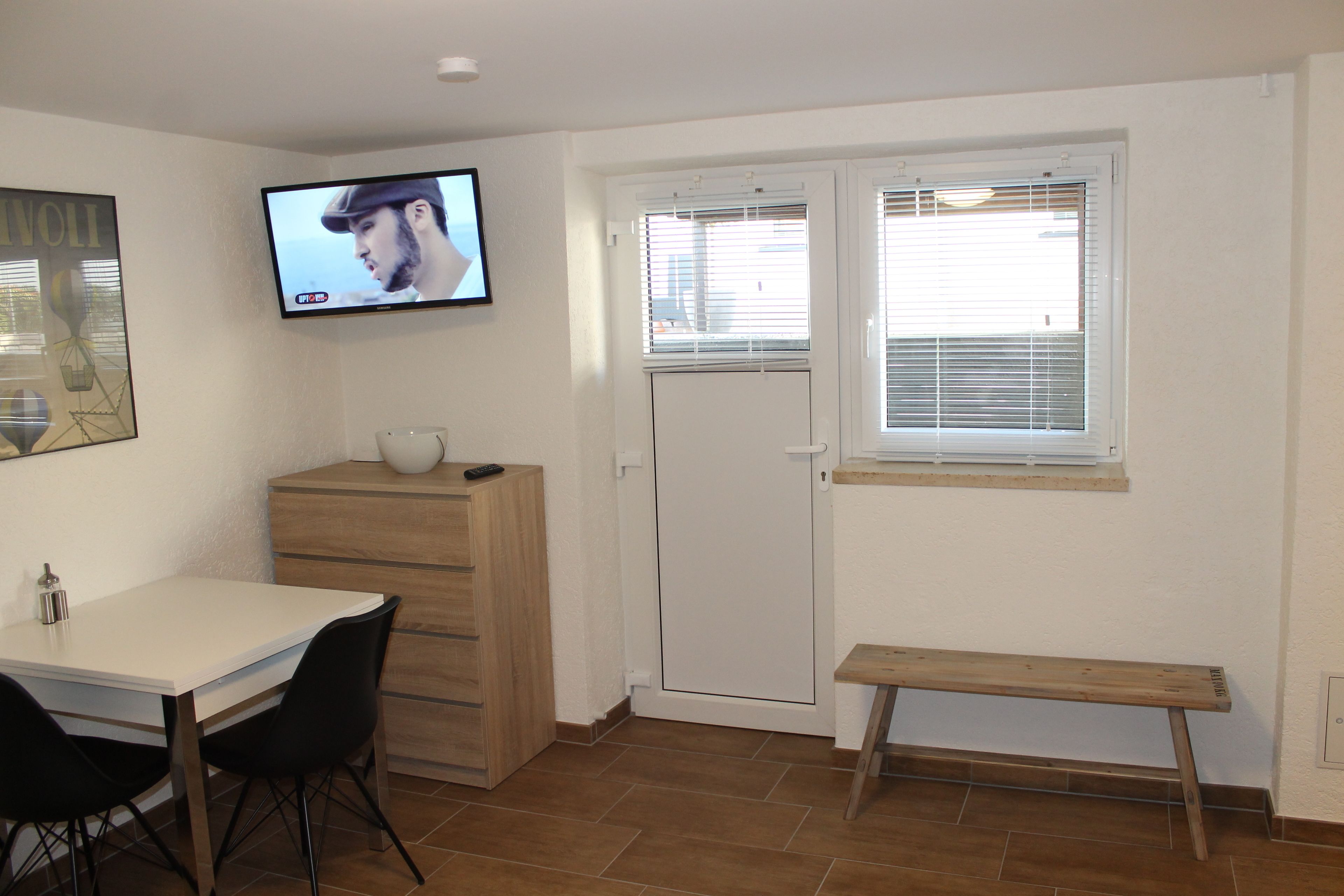 A room with a white table, two black chairs, a wooden bench, a TV on the wall, a door with a window, and a cabinet.